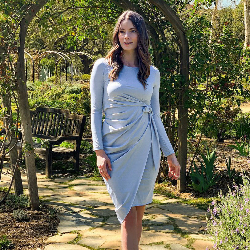 Woman in a light blue-gray dress standing in a garden with trees and a bench in the background.